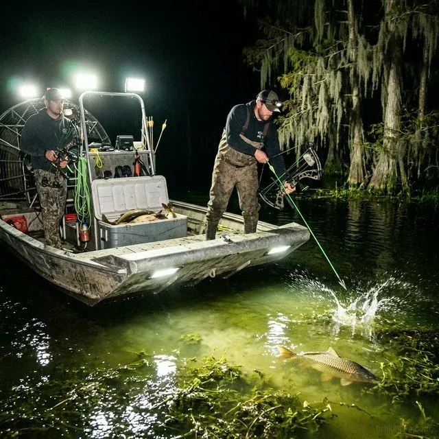 Two bowfishermen on an airboat at night with LED floodlights, arrow splashing into water targeting carp in a Southern swamp