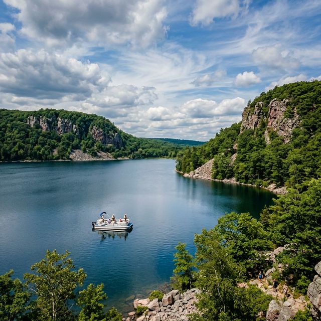 Panoramic view of Devil's Lake in Wisconsin's Baraboo Range during summer, with dramatic quartzite bluffs rising 500 feet above the pristine lake and a pontoon boat visible on the calm water surrounded by lush hardwood forest